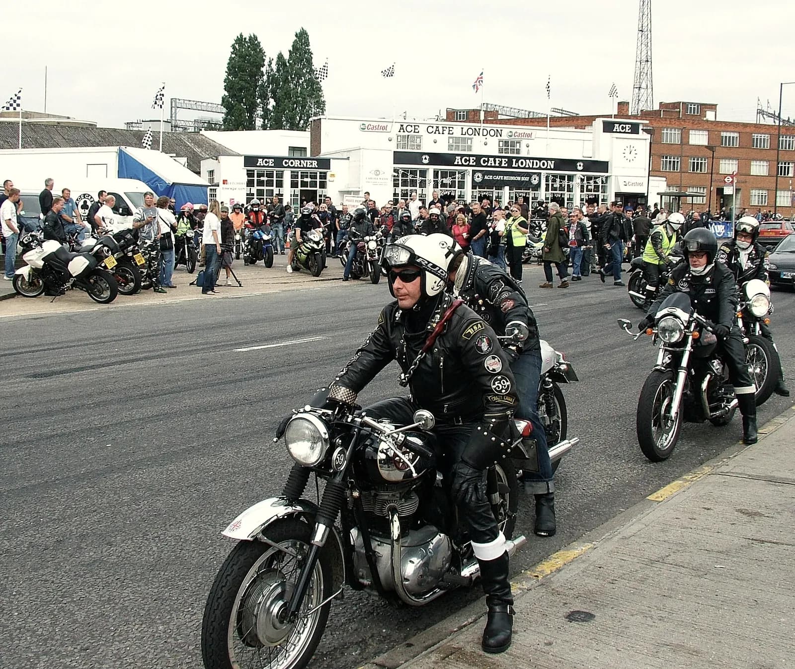 Leather-jacketed riders on vintage British motorcycles pulling out from the Ace Cafe in London