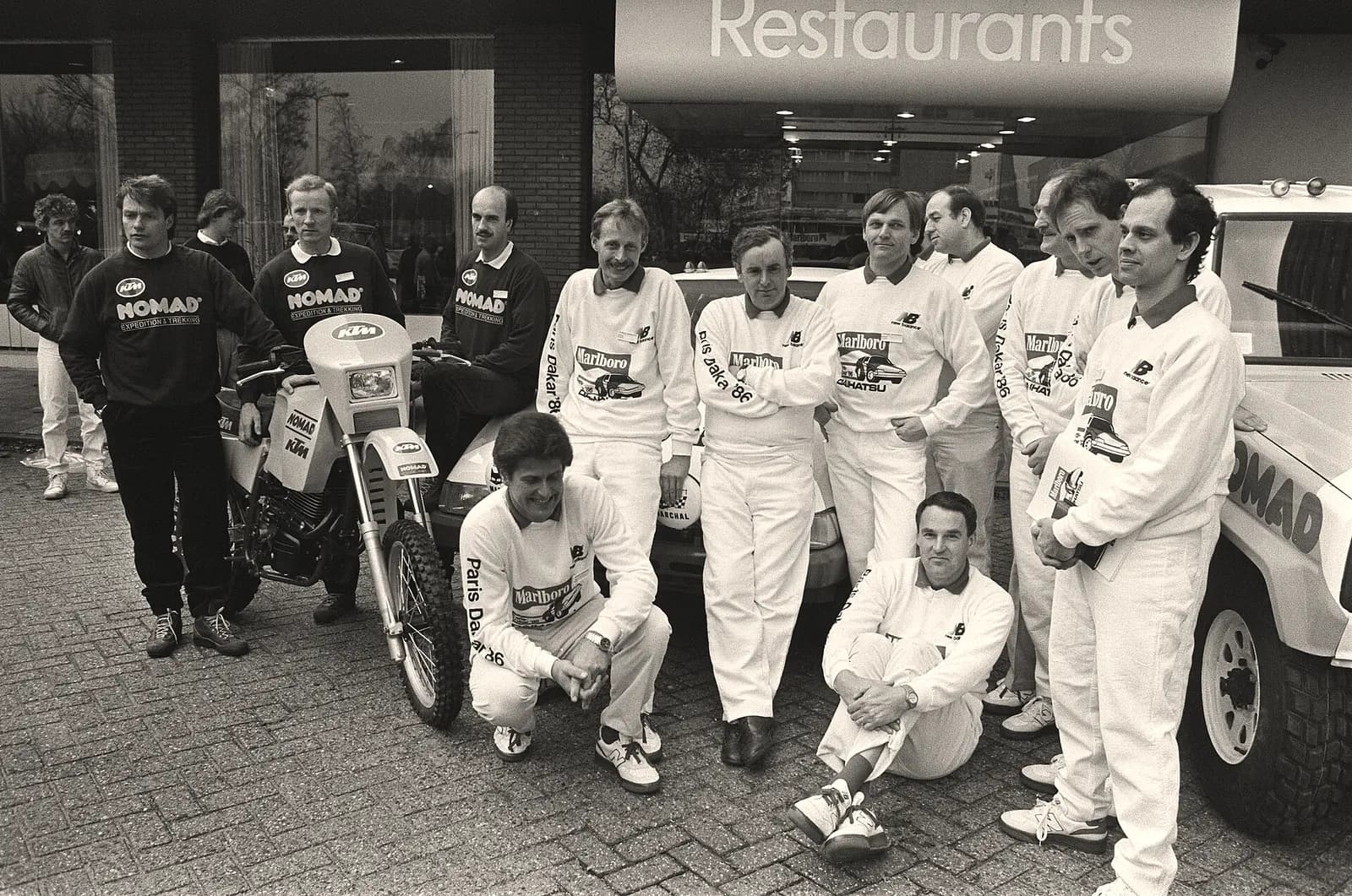 Black and white photo of a 1985 Paris-Dakar rally team posing in white race suits beside a motorcycle