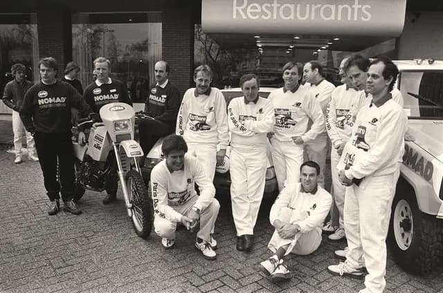 Black and white photo of a 1985 Paris-Dakar rally team posing in white race suits beside a motorcycle